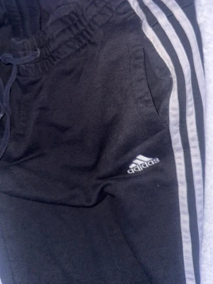 Svarta Adidas mjukisbyxor med ränder - Säljer ett par svarta mjukisbyxor från Adidas med klassiska vita ränder längs benen och logga på låret. Byxorna har snörning i midjan och sidofickor. Perfekta för chill eller träning.