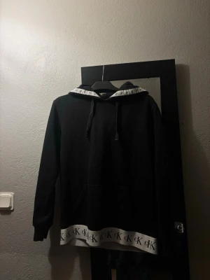 Svart hoodie från Calvin Klein - Svart hoodie med huva och dragsnören från Calvin Klein. Vit kant med CK-logga längs nederkanten och insidan av huvan. Klassisk känguruficka framtill och långa ärmar. Perfekt för en chill och stilren look.