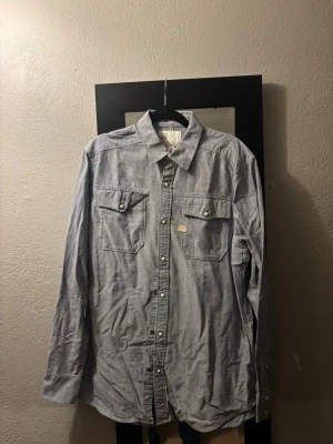 Blåvit randig skjorta med knappar - Säljer en blåvit randig skjorta med långa ärmar och klassisk krage. Skjortan har två bröstfickor med lock och knappar framtill. Perfekt för en avslappnad men ändå stilren look.