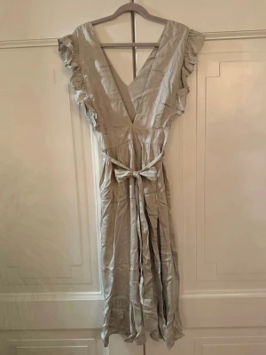 Långklänning med volang - Snygg långklänning med v-ringning, volangdetaljer vid ärmarna och ett matchande knytband i midjan. Klänningen har en omlott-look, slits och dragkedja i sidan. Tillverkad i ett lätt tyg som passar perfekt för varmare dagar. Från kollektionen nakd x Linn Ahlborg