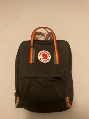 Svart Kånken ryggsäck med regnbågshandtag - Klassisk svart Fjällräven Kånken ryggsäck med regnbågsfärgade handtag och detaljer. Ryggsäcken har en rund röd logotyp framtill, stort huvudfack och en mindre ficka med dragkedja på framsidan. Perfekt för skola eller vardag.