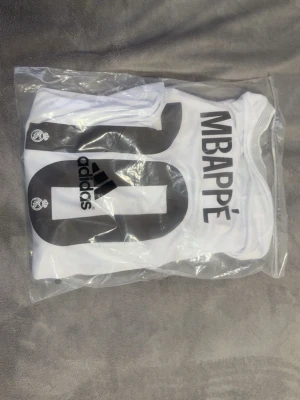 Vit Real Madrid tröja Adidas - Säljer en vit Real Madrid matchtröja från Adidas med svart tryck, nummer 20 och namnet Mbappé på ryggen. Tröjan har klubbens logga och Adidas-loggan tryckt på ryggen. Perfekt för dig som älskar fotboll och vill ha en snygg supportertröja. Helt ny 