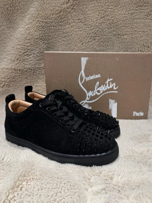 Svarta Christian Louboutin sneakers - Säljer ett par svarta sneakers från Christian Louboutin med svarta nitar på tån och klassisk röd sula. Skorna har snörning, rund tå och är tillverkade i mocka med läderdetaljer. Perfekta för dig som vill sticka ut med en lyxig och edgy stil.