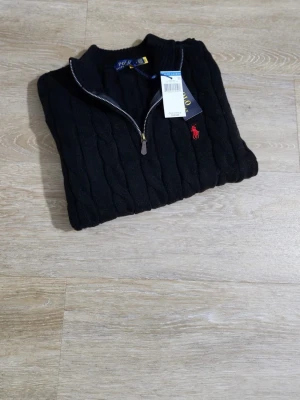 Svart stickad half zip från Polo Ralph Lauren - Kabelstickad svart tröja från Polo Ralph Lauren med halv dragkedja och klassisk röd logga broderad på bröstet. Tröjan har ribbade muddar och hög krage, perfekt för kyliga dagar. Snygg och tidlös design som funkar till allt.