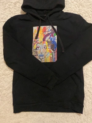 Svart hoodie med färgglatt konsttryck - Cool svart hoodie med huva och dragsko. På bröstet finns ett stort färgglatt konsttryck med ansikte och abstrakta former i blått, rött, gult och orange. Perfekt för dig som gillar unika plagg med streetkänsla.