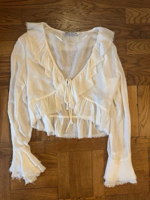 Vit volangblus med knytning - Söt vit blus med volangdetaljer och knytning framtill. Blusen har djup v-ringning, långa ärmar och råa kanter för en avslappnad look. Tillverkad i ett tunt, luftigt tyg som ger en bohemisk känsla.