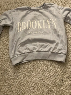 Ljusblå sweatshirt med BROOKLYN-tryck - Ljusblå sweatshirt med stort vitt BROOKLYN-tryck och texten NEW YORK CITY på bröstet. Tröjan har rund halsringning, ribbade muddar vid ärmslut och nederkant samt en avslappnad passform. Perfekt för en casual streetwear-look.