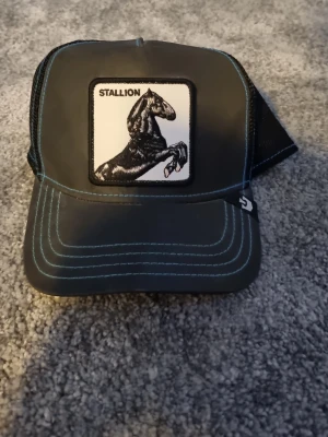 Svart keps Goorin Bros Stallion - Snygg svart truckerkeps från Goorin Bros med broderad patch framtill som visar en stegrande häst och texten 'STALLION'. Kepsen har mesh baktill för bra ventilation och blå kontrastsömmar. Justerbar storlek med snapback köptes för 700 nästan ny använt 2 ggr
