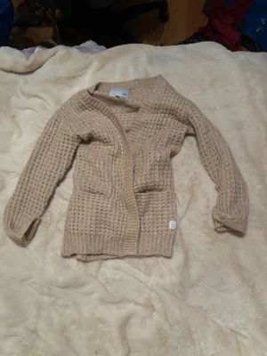 Beige stickad kofta från Name It - Mysig beige stickad kofta från cubus med grovstickat mönster och två framfickor. Koftan har långa ärmar och ribbade muddar vid ärmslut och nederkant. Perfekt för lager på lager och enkel att matcha med olika outfits.