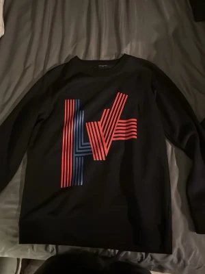 Svart Louis Vuitton sweatshirt - Säljer en svart sweatshirt från Louis Vuitton med ett stort grafiskt LV-tryck i rött och blått på framsidan. Tröjan har rund halsringning och långa ärmar. Säljer den just för att den inte passar mig eftersom jag är 190cm och den blir lite för kort på midjan. Passar perfekt för 165-185