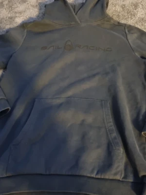 Mörkblå hoodie från Sail Racing - Snygg mörkblå hoodie från Sail Racing med broderad logga på bröstet. Klassisk känguruficka framtill och stor huva. Perfekt för en avslappnad och sportig stil. Tillverkad i mjukt bomullsmaterial för extra komfort.