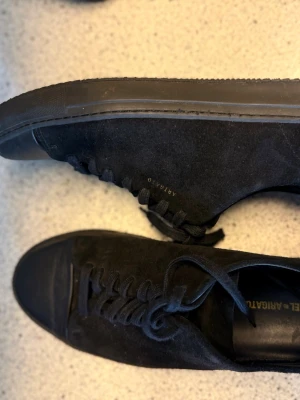 Svarta sneakers från Axel Arigato - Säljer ett par stilrena svarta sneakers från Axel Arigato i mocka med diskret guldfärgad logga på sidan. Skorna har rund tå, platt sula och klassisk snörning. Perfekta för dig som gillar minimalistisk design och hög kvalitet.