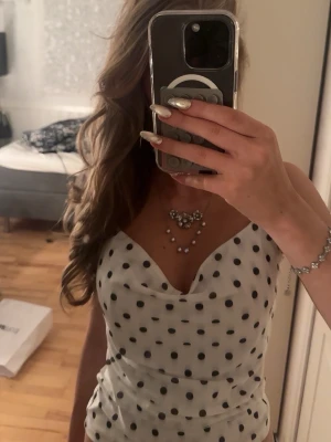 Vit prickig omlottopp - Snygg vit omlottopp med svarta prickar och draperad urringning. Toppen har ärmlös design och figurnära passform, perfekt för att lyfta din stil. Materialet känns lätt och svalt, vilket gör den skön att bära under varma dagar.