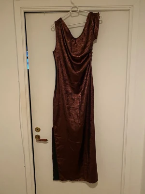 Lång brun glittrig klänning - Säljer en långklänning i skimrande brun färg med glittrig finish. Klänningen har en draperad urringning och är ärmlös, vilket ger en elegant och trendig look. Perfekt för dig som vill sticka ut på nästa event!