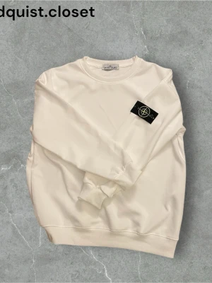 Vit tröja från Stone Island - Vit sweatshirt från Stone Island med klassisk rund halsringning och ribbade muddar. Tröjan har den ikoniska svarta logotypen med kompass på vänster ärm. Tillverkad i mjuk bomull för en clean och stilren look. Det står storlek M men den är ganska liten i storlek så passar bättre som storlek S.