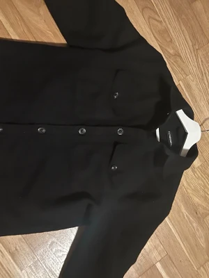 Svart overshirt från J.Lindeberg - Säljer en svart overshirt från J.Lindeberg med klassisk krage, två bröstfickor med knapp och raka ärmar. Jackan har tryckknappar längs hela framsidan och är gjord i ett mjukt ullmaterial. Perfekt för lager på lager och stilren look.
