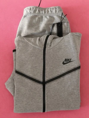 Nike tech fleece set - Snygga ljusgrå mjukisbyxor från Nike med svarta dragkedjor på fickorna och elastisk midja med snörning. Perfekta för chill eller träning, med klassisk Nike-logga och mjukt material som känns skönt mot huden. Säljer koftan och byxorna för endast 1000kr!! Några frågor ? Fråga helst! Nike logot på byxorna har gått bort lite eftersom av tvättningen, ett av snören är också lite sönder. Fråga bara efter bilder!