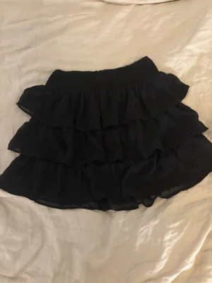 Svart volangkjol med resårmidja - Supersöt svart kortkjol med flera lager volanger och bred resår i midjan. Kjolen är luftig och har en flowy känsla, perfekt till sommaren eller våren. Materialet känns lätt och mjukt mot huden. Lappen är bort så kommer inte ihåg var den är ifrån men förmodligen typ HM, Lindex, Gina mm