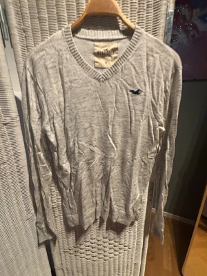 Ljusgrå stickad tröja Hollister - Snygg ljusgrå stickad tröja från Hollister med V-ringning och diskret mörkblå logga på bröstet. Tröjan har långa ärmar och ribbade muddar vid ärmslut och nederkant. Perfekt för lager på lager och enkel att matcha.