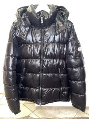 Svart Moncler dunjacka med huva - Glansig svart dunjacka från Moncler med quiltad design och avtagbar huva. Jackan har dragkedja framtill, två sidofickor med dragkedja och klassisk Moncler-logga på ärmen. Perfekt för kalla vinterdagar och ger en clean, modern look.