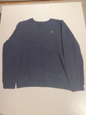 Blå sweatshirt från Lyle & Scott - Mörkblå sweatshirt från Lyle & Scott Junior med klassisk rund hals och gul broderad logga på bröstet. Tröjan har långa ärmar och ribbade muddar vid ärmslut och nederkant. Perfekt för en chill och stilren look.