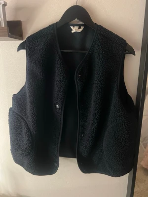 Svart teddyväst med tryckknappar - Mysig svart väst i teddy-material med rund halsringning och stora fickor framtill. Västen har tryckknappar och en avslappnad passform, perfekt att slänga över en hoodie eller tröja för extra värme. Enkel och stilren design utan synligt märke.