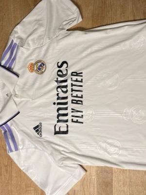 Real Madrid matchtröja Adidas vit - Snygg vit Real Madrid matchtröja från Adidas med klubbmärke och sponsortryck på bröstet. Tröjan har lila detaljer på axlarna, svart Adidas-logga och klassisk krage med knapp. Tillverkad i lätt och ventilerande material, perfekt för fotboll eller träning.