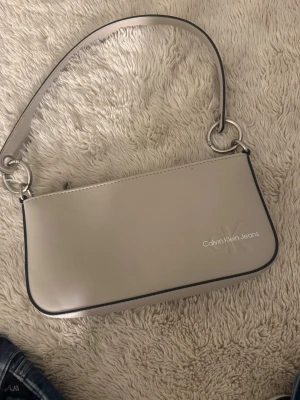 Beige axelremsväska Calvin Klein Jeans - Snygg beige axelremsväska från Calvin Klein Jeans i stilren design. Väskan har silverfärgade metalldetaljer, dragkedja upptill och logga på framsidan. Tillverkad i slätt konstläder och har en smidig, rektangulär form.
