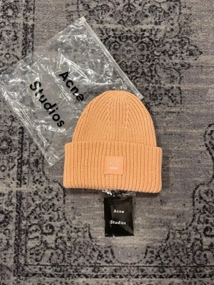 mössa Acne Studios - Säljer en beige ribbstickad mössa från Acne Studios med uppvikt kant och det klassiska Face-logotypen framtill. Mössan är i mjukt material och har en clean, modern look som passar perfekt till streetwear och casual outfits.