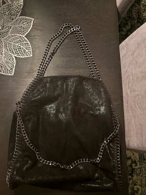 Svart axelväska med kedjedetaljer - Snygg svart axelväska med glansig finish och silverfärgade kedjor som axelband och runt kanterna. Väskan har en mjuk form och innerfack med dragkedja. Perfekt för dig som gillar en edgy och trendig look.