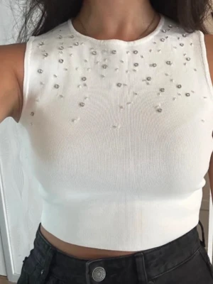 Vit croppad linnetopp med pärlor - Säljer en vit croppad linnetopp med rund hals och ärmlös design. Toppen är dekorerad med små pärlor framtill som ger en snygg detalj. Ribbad struktur i materialet och tight passform som framhäver siluetten. Perfekt till höga jeans eller kjol. Står M men passar S.
