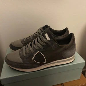 Grå sneakers från Philippe Model - Snygga grå sneakers från Philippe Model med coola detaljer i mocka och mesh. Skorna har en vit och svart randig sula, snörning och den klassiska skölden på sidan. Perfekta för dig som gillar stilrena. Lite slitna vid sulan men det är inget man tänker på.
