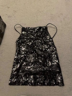 Svart paljettlinne med smala band - Säljer ett svart linne från H&M täckt med glittriga paljetter. Toppen har smala justerbara axelband och rak halsringning. Perfekt för dig som vill sticka ut och glänsa lite extra. Insidan är fodrad i mjuk polyester för skön känsla.