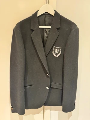 Svart kavaj från Tiger of Sweden - Snygg svart kavaj från Tiger of Sweden med emblem på bröstet. Klassisk modell med två knappar framtill och fickor. Materialmix av akryl, polyester och ull. Perfekt för dig som vill ha en stilren och tidlös look.