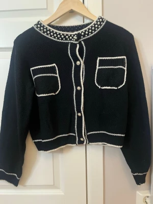 Svart kofta med pärldetaljer - Säljer en svart stickad kofta med vita kanter och dekorativa pärlor runt halsen