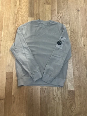  sweatshirt från C.P. Company - Snygg sand grå sweatshirt från C.P. Company med deras ikoniska linsdetalj på ärmen och en liten ficka med knapp. Tröjan har rund halsringning, ribbade muddar och är tillverkad i mjukt bomullsmaterial. Perfekt för en avslappnad och trendig stil. Obs priset ej hugget i sten. 