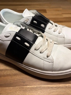 Valentino Garavani vita sneakers - Snygga vita sneakers från Valentino Garavani med bred svart detalj över sidan och vita snören. Skorna har rund tå och platt sula, samt guldfärgad logga på plösen. Tillverkade i skinn för en exklusiv känsla.