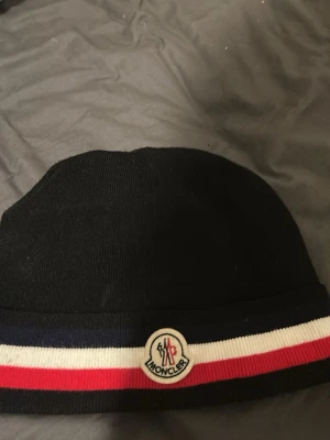 Svart Moncler mössa med randig kant - Snygg svart mössa från Moncler med ribbad kant i blått, vitt och rött. Framtill sitter Monclers klassiska logotyp. Mössan är stickad och har en stilren, enkel form som passar perfekt till höst och vinter.