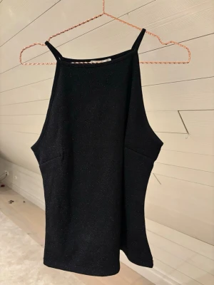 Svart glittrig linnetopp - Snygg svart linnetopp med tunna axelband och glittrig. Toppen har en rak, något loose passform och hög halsringning.