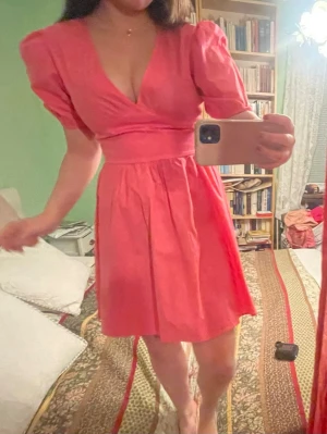 Rosa kortklänning med puffärm - Söt rosa kortklänning med v-ringning och puffärmar. Klänningen har markerad midja och lätt utställd kjol som ger en feminin siluett. Perfekt för sommarens alla tillfällen. Materialet känns mjukt och lätt, troligen bomull eller polyester.