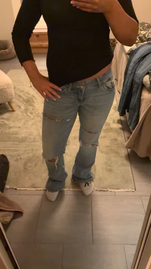 Ljusblå slitna bootcut jeans - Säljer ett par ljusblå jeans med slitningar och hål på benen. Modellen har bootcut passform och normal midja. Jeansen är tillverkade i klassiskt denimtyg och har fem fickor samt knappgylf. Perfekta för en avslappnad och trendig look.