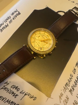 Michael Kors guldfärgad klocka - Stilren klocka från Michael Kors med guldfärgat ur och gnistrande stenar runt boetten. Urtavlan har flera små visare och bruna läderband med klassiskt spänne. Perfekt accessoar för dig som gillar en lyxig och elegant look.