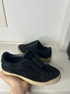 Svarta mocka slip-on sneakers - Svarta slip-on sneakers i mocka med vit sula och minimalistisk design. Skorna har elastiska paneler på ovansidan för enkel påtagning och en klassisk rund tå. Perfekta för en avslappnad och stilren look.