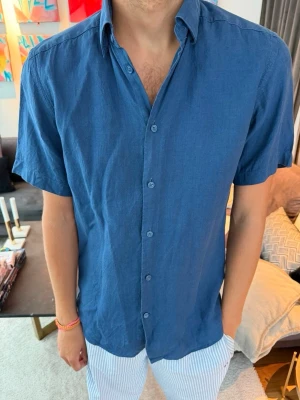 Blå kortärmad skjorta - Snygg blå skjorta med korta ärmar och klassisk krage. Skjortan har knappar framtill och en avslappnad passform, perfekt för en stilren och bekväm look.