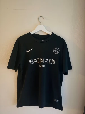 Svart Balmain x PSG t-shirt - Snygg svart t-shirt från Balmain i samarbete med PSG och Nike. T-shirten har klassisk passform, rund hals och korta ärmar. Stor Balmain Paris-logga framtill samt PSG och Nike-loggor. Perfekt för dig som gillar streetwear och sportig stil.