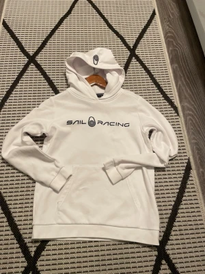 Vit hoodie från Sail Racing - Snygg vit hoodie från Sail Racing med svart logga och text på bröstet. Klassisk känguruficka framtill och huva med tryckt logga. Tillverkad i mjukt material som känns skönt mot huden. Perfekt för en avslappnad och sportig stil.
