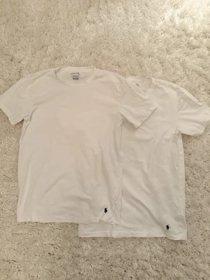 Vita t-shirts från Polo Ralph Lauren - Två klassiska vita t-shirts sopm har en liten broderad logga i svart vid nederkanten. T-shirtsen har rund halsringning och korta ärmar. Perfekta basplagg från Polo Ralph Lauren i mjuk bomull.