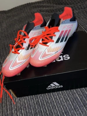 Adidas F50 fotbollsskor vit/röd - Säljer ett par Adidas F50 fotbollsskor i vitt och rött med svarta detaljer och klassiska tre ränder på sidan. Skorna har snörning och är tillverkade i syntetmaterial för lätt känsla på planen. Snygg design med neonröda skosnören och detaljer.