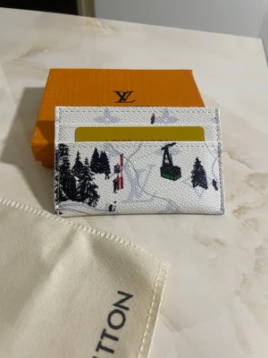 Vit korthållare - Vit korthållare med vinterinspirerat tryck som visar granar, skidåkare och en lift. Plånboken har plats för kort och är tillverkad i tåligt skinn. Perfekt för dig som vill ha något unikt och stilrent i fickan.