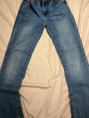 Blå jeans med papperspåsemidja från 157 - Säljer ett par ljusblå jeans från 157 med papperspåsemidja och klassisk femficksdesign. Jeansen har raka ben och är tillverkade i bomullsmaterial med snygga kontrastsömmar. Perfekta för en avslappnad och trendig look.